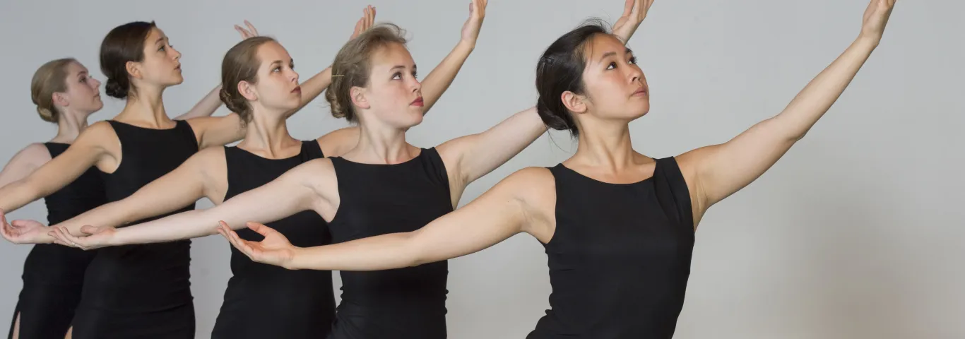 a line of five women in black dresses with arms held out in a ballet position