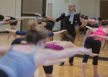 A woman wearing a black body suit and scarf, leads a crowded room of dancers